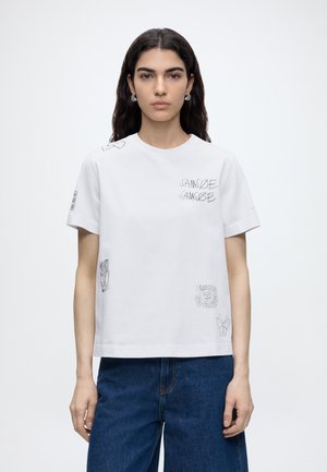 Junge Frau mit langen dunklen Haaren, die ein weißes T-Shirt mit kurzen Ärmeln und kleinen schwarzen grafischen Motiven sowie blaue Jeans trägt, steht frontal.
