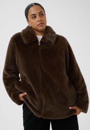 Femme portant une veste zippée en fausse fourrure marron, les mains dans les poches, regardant de côté sur un fond uni.