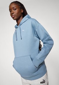 Light blue hoodie with a kangaroo pocket, drawstring, and white logo on the chest. Soft fabric and relaxed fit visible. Grey sweatpants below.