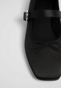 Chaussure plate Mary Jane noire avec un bout carré, un fin détail de nœud et une bride à boucle argentée sur fond clair.