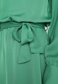 Grüne Bluse mit glatter Textur, ausgestattet mit einem gebundenen Taillenband und plissiertem Detail. Lange Ärmel mit gerafften Manschetten, die eine lässige Passform verleihen.