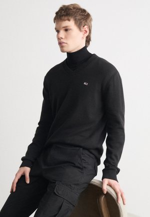 Suéter negro de cuello en V con puños y cuello acanalados, combinado con pantalones cargo negros. Pequeño logotipo en el área del lado izquierdo del pecho.