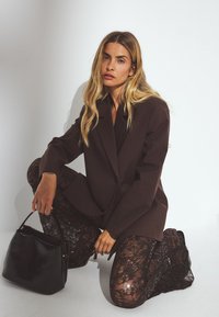 Bruine oversized blazer, doorzichtige zwarte kant broeken en een zwarte handtas. De blazer heeft een enkele knoop en de broeken hebben ingewikkelde kantpatronen.