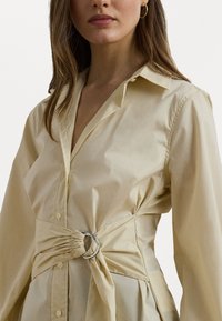 Blusa amarelo claro com colarinho estruturado, frente com botões, mangas longas e cintura ajustada com um detalhe em anel prateado.