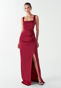 Ženska v brezrokavnem rdečem večernem obleku s kvadratnim izrezom in visokim razporkom, stoječa pred enobarvno belo ozadjem.