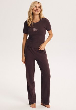 Frau steht barfuß, trägt ein dunkelviolettes, geripptes Kurzarmshirt und passende weit geschnittene Hose mit dem Text "French Stories" auf dem Shirt.