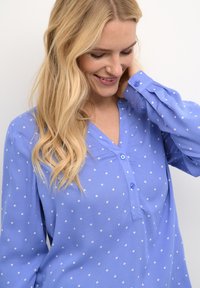Blouse à manches longues bleu clair avec des pois blancs, décolleté en V et détails de boutons. Confectionnée dans un tissu doux avec une coupe décontractée.