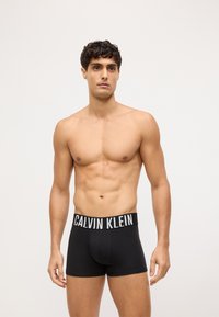 Czarne bawełniane bokserki z białym paskiem z logo "CALVIN KLEIN", charakteryzujące się dopasowanym krojem i gładką teksturą. Model stoi z atletyczną sylwetką.