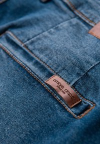 Tissu en denim bleu foncé avec une étiquette en cuir marron portant l'inscription "ombre dept." Couture visible et surface texturée.