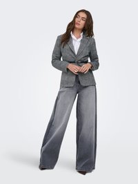 Mujer con blazer gris con cinturón negro sobre una camisa blanca y vaqueros grises de pierna ancha y descoloridos, de pie con las manos juntadas.