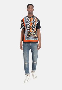 T-shirt multicolore a maniche corte con un bold pattern, dettagli arancioni e maniche nere, abbinato a un paio di jeans blu strappati e sneakers bianche.