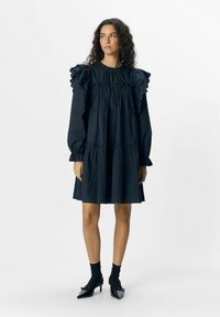 Navyblaues Kleid mit gerafften Ärmeln, gestuftem Design und lockerer Passform. Glatte Textur und knielang, kombiniert mit schwarzen Socken und spitzen Schuhen.