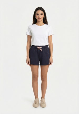 Femme debout, de face, portant un t-shirt blanc à manches courtes, un short bleu marine avec un cordon rose et des baskets beige.