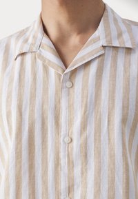 Camisa a rayas con un patrón en beige claro y blanco, hecha de una tela texturizada, con cuello en pico y cierre de botones en la parte delantera.
