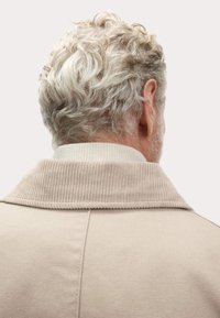 Beige jas met een geribbelde kraag, gezien vanuit de achterkant. Gestructureerde stof en golvend lichtgekleurde haar zichtbaar boven de kraag.