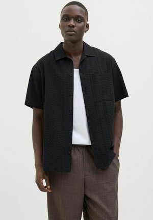 Camisa negra de manga corta con un diseño texturizado, cierre de botones y bolsillo frontal, llevada sobre una camiseta blanca y combinada con pantalones de cuadros.