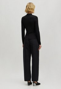 Schwarzes langärmeliges Oberteil mit gerippter Textur und hohem Kragen. Kombiniert mit locker sitzenden dunklen Hosen und schwarzen Schuhen, von hinten betrachtet.