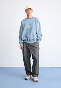 Lyseblå genser med ribbestrikkede ermer og nederkant, grå bukser og hvite joggesko med beige detaljer. Modellen har på seg en beige caps.