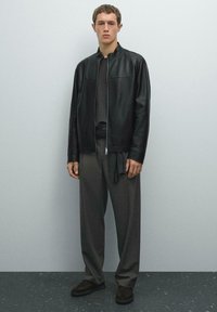 Young man standing against a gray wall wearing a black zip-up leather jacket, dark gray wide-leg pants, and black shoes.
