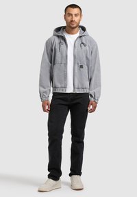 Homme debout portant un sweat à capuche gris zippé, un T-shirt blanc, un jean noir et des baskets blanches sur un fond gris clair.