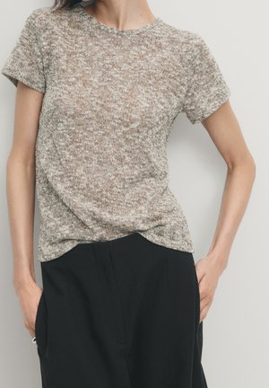 Donna che indossa un top a maniche corte, beige e grigio, lavorato a maglia, infilato in pantaloni neri a vita alta con le mani nelle tasche.