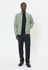 Világoszöld bomberdzseki fekete pólóval, sötét farmerrel és fekete cipővel. A dzseki sima textúrájú és bordázott mandzsettákkal rendelkezik.