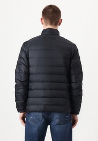 Schwarze Steppjacke mit horizontalem Steppmuster, hohem Kragen und dünner Stoffstruktur. Getragen über blauen Jeans.