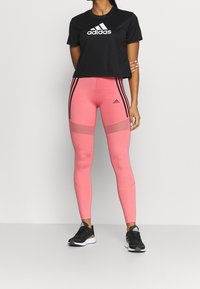 Černé zkrácené tričko s bílým logem "adidas", doplněné růžovými legínami s černými pruhy a síťovanými panely. Černé sportovn í boty.