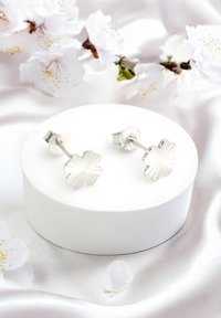 Silberne blumenförmige Steckerohrringe mit linearen Mustern, auf einem glatten weißen Display montiert, vor einem weichen, hellen Stoffhintergrund.