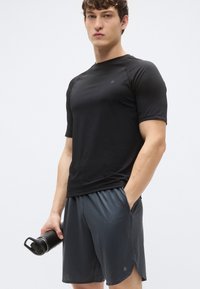 Tricou sport negru cu mâneci scurte, asortat cu pantaloni scurți gri închis, ambele realizate dintr-un material ușor. Modelul ține în mână o sticlă de apă neagră.