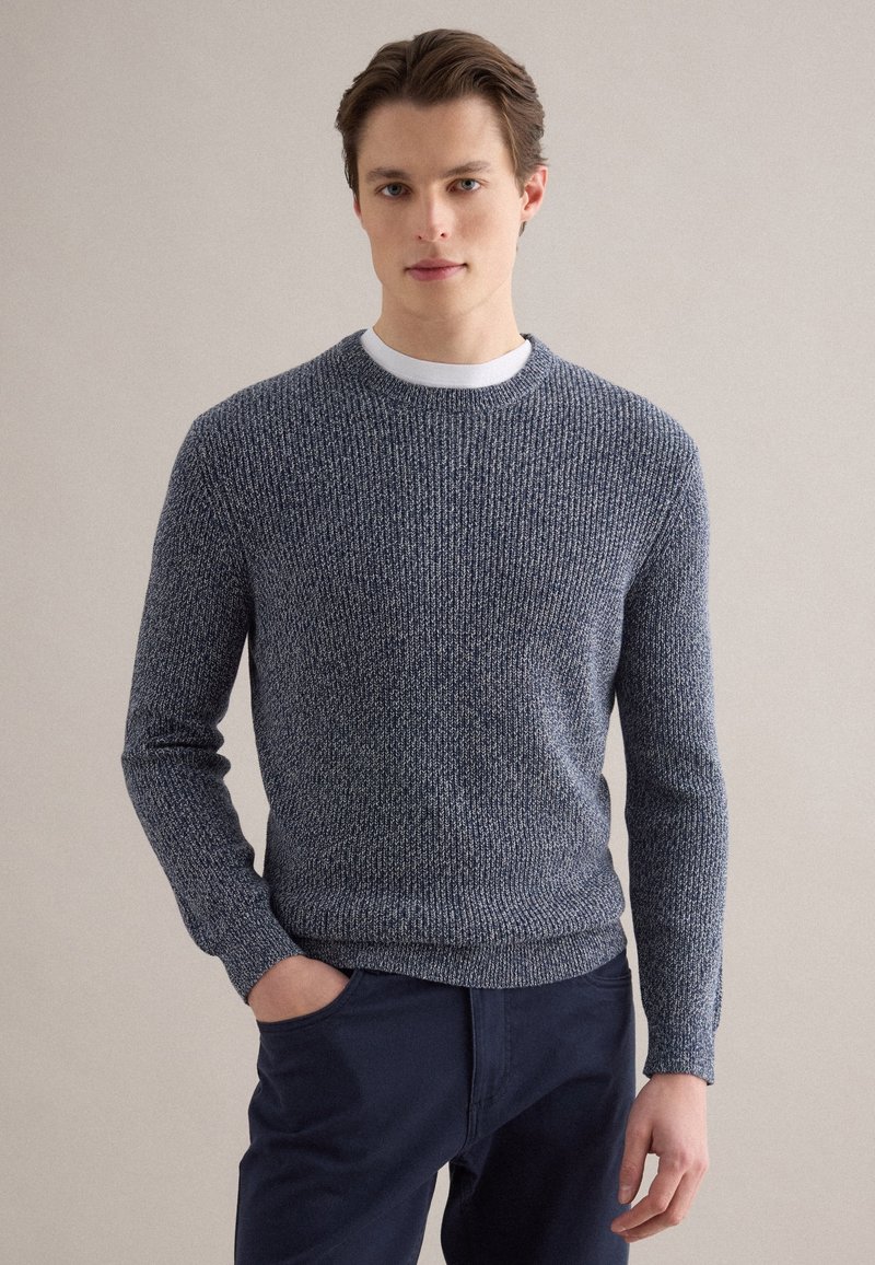 Junger Mann, der einen blauen strukturierten Strickpullover über einem weißen Hemd mit marineblauen Hosen trägt, steht mit einer Hand in der Tasche vor einem neutralen Hintergrund.