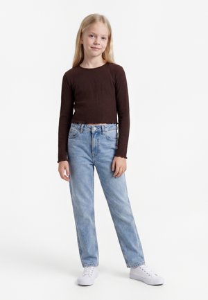 Jeune fille aux longs cheveux blonds portant un haut marron foncé côtelé à manches longues, un jean bleu clair et des baskets blanches, debout sur un fond blanc.