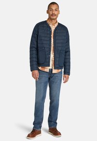 Chaqueta acolchada de color marino con botones a presión, bolsillos frontales y cuello redondeado, superpuesta a una camisa de cuadros, combinada con jeans azules y botas marrones.
