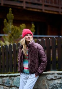 Hnedá prešívaná bunda s chlpatou textúrou, ružová pletená čiapka a viacfarebný pruhovaný sveter; stojí vedľa dreveného plota.