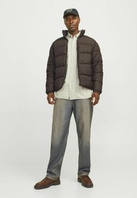 Chaqueta acolchada negra sobre una camisa de cuadros verdes, con unos vaqueros ligeros de color azul claro y zapatos de cuero marrón, complementada con una gorra oscura.
