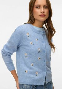 Kobieta ubrana w jasnoniebieski rozpinany kardigan z haftowanymi małymi białymi i żółtymi kwiatami, w połączeniu z dżinsami.