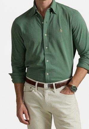 Hombre con camisa verde abotonada con mangas remangadas, pantalones beige, cinturón marrón y reloj de pulsera con esfera verde y correa marrón.