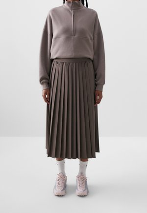 Personne portant un pull taupe à zip quart, une jupe midi plissée assortie, des chaussettes blanches Nike et des baskets roses et grises, debout devant un fond uni.