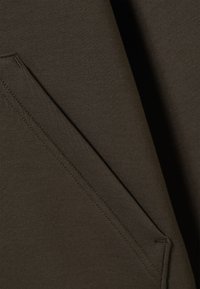 Dunkelolivgrüner Stoff mit glatter Textur, der eine diagonale Tasche mit sichtbaren Nähten aufweist. Einfaches, minimalistisches Design.