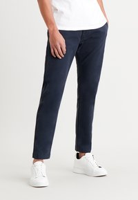 Pantalon chino bleu marine en sergé de coton, avec une coupe droite, des poches latérales et des détails cousus le long des jambes, porté avec des baskets blanches.