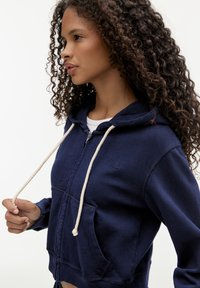 Felpa con zip blu navy in tessuto morbido, dotata di tasca a marsupio, cordini crema e logo ricamato sul petto.