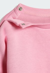 Ružový sveter s jemnou textúrou, ktorý má zapínanie na cvoky na ramene. Tkanina má hladký povrch a je vyrobená z bavlnenej zmesi.