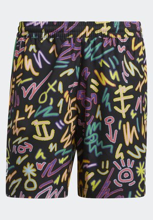 Henkilö, jolla on värikkäät shortsit, joissa on abstrakteja symboleja, valkoiset sukat mustilla raidoilla ja siniset Adidas-tossut, seisoo yksinkertaisen taustan edessä.