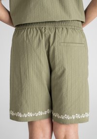 Olivengrønne shorts med lodrette striber, elastisk talje og baglomme; prydet med hvid blomsterbroderi ved kanten.