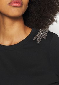 Svart t-shirt med rund halsringning som har en texturerad dekoration på vänster axel bestående av mörkfärgade pärlor i ett bladmönster.