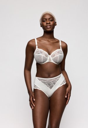 Jonge zwarte vrouw met kort blond haar die een witte kanten bh en bijpassende hoogwateronderbroek modelleert tegen een effen lichte achtergrond.