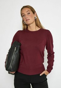Maglione in maglia bordeaux con scollatura rotonda, maniche lunghe e orlo a coste; abbinato a una borsa in pelle nera con dettagli a zip.