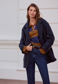Marineblauwe jas met gouden knopen, bruin-blauw gestreepte trui, donker denim broek en een bruine riem. Model staat voor een neutrale achtergrond.