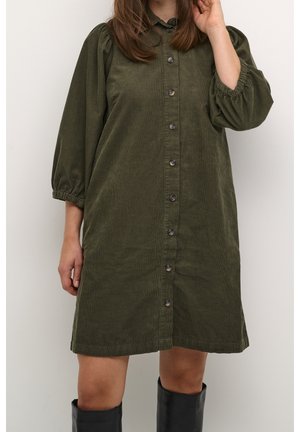 Femme portant une robe boutonnée en velours côtelé vert olive longueur genou avec des manches bouffantes et des bottes noires montantes, debout contre un mur blanc.