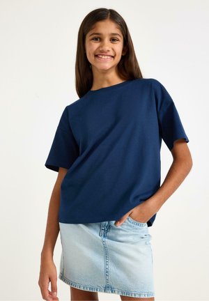 Smilende pige med langt mørkt hår iført en mørkeblå T-shirt og en lyseblå denimnederdel, stående med den ene hånd i lommen.